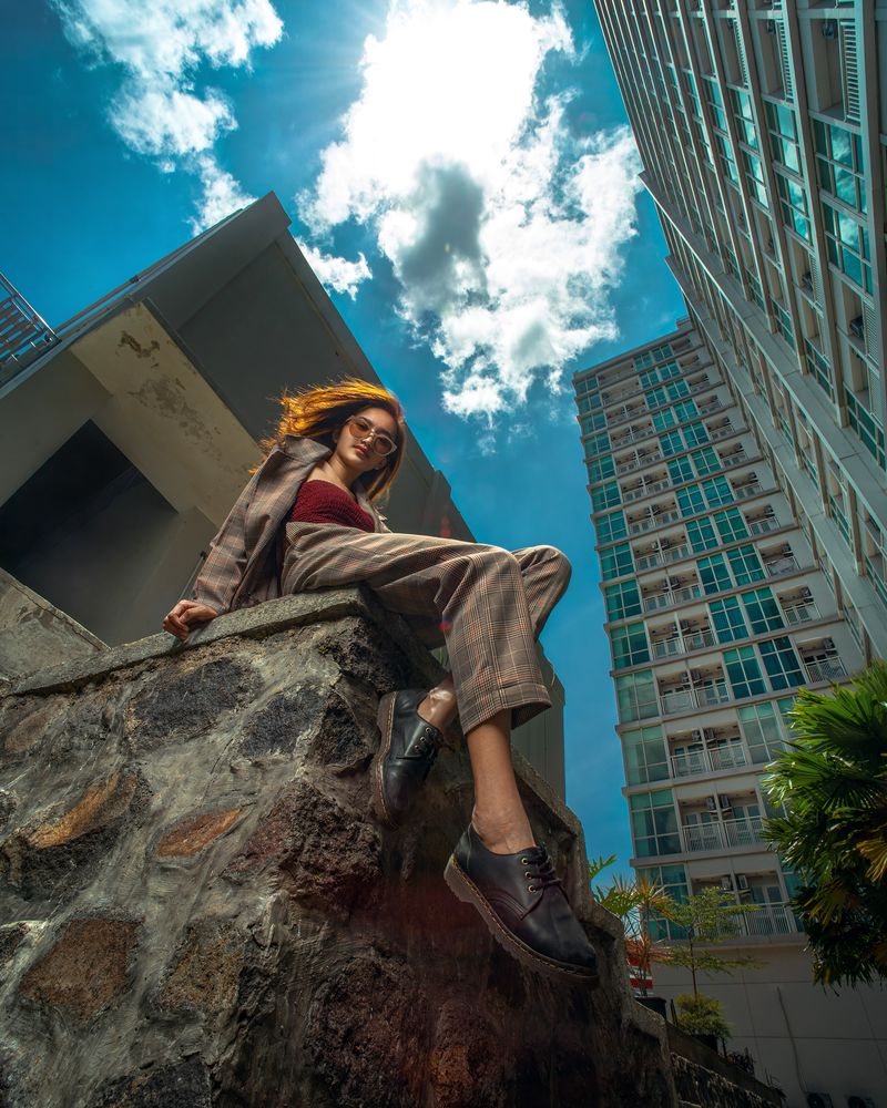 Low-angle creative portrait of a woman seated on a stone wall beneath city buildings and a blue sky