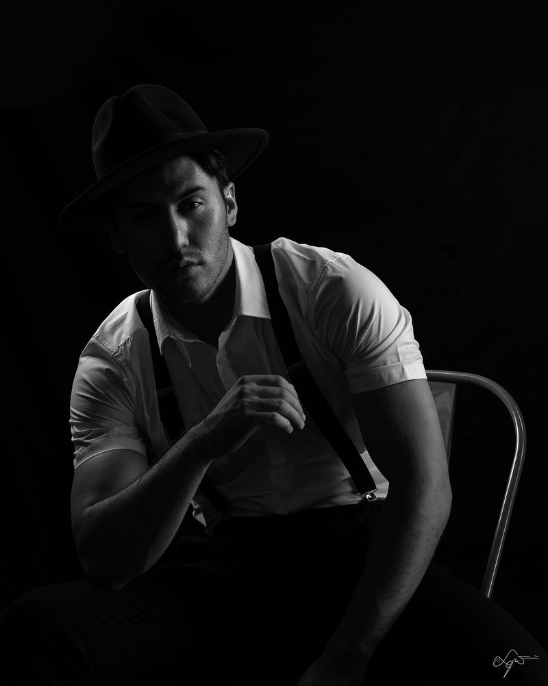 Black-and-white creative portrait of a seated man in a hat, white shirt, and suspenders
