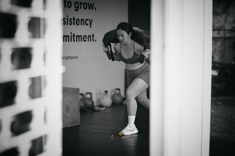 Black-and-white view of an athlete carrying a sandbag, framed through gym equipment